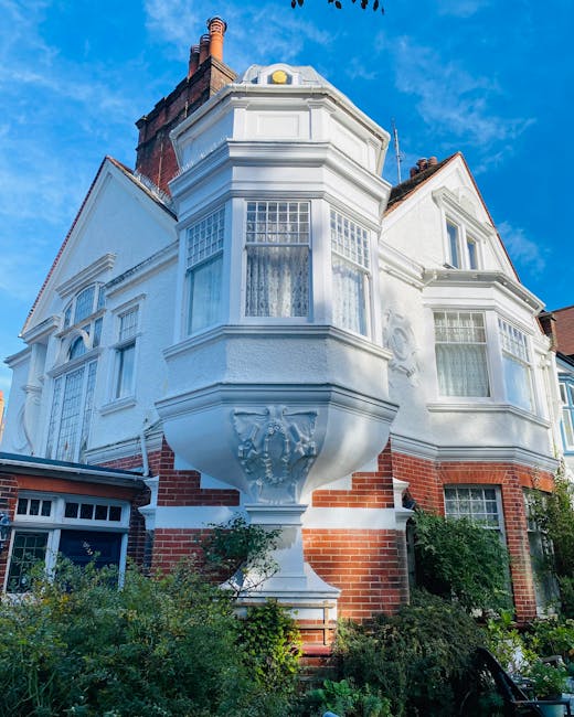 The exterior of a Victorian-style house in Shirley with white weatherboarding and large bay windows, part of a home undergoing furniture transport and packing for a house removal. In the foreground, green shrubs and plants are visible along the base of the building. Exterior walls include brickwork at the lower level and decorative woodwork and detailed cornices around the bay windows. The clear blue sky provides natural light, illuminating the façade. This setting suggests active packing or preparation for a home relocation, with the focus on safeguarding the property during furniture loading processes. Man with Van Shirley, a professional removals service, routinely arranges careful loading of furniture and boxes to prevent damage, especially when dealing with historic Victorian houses like this one.