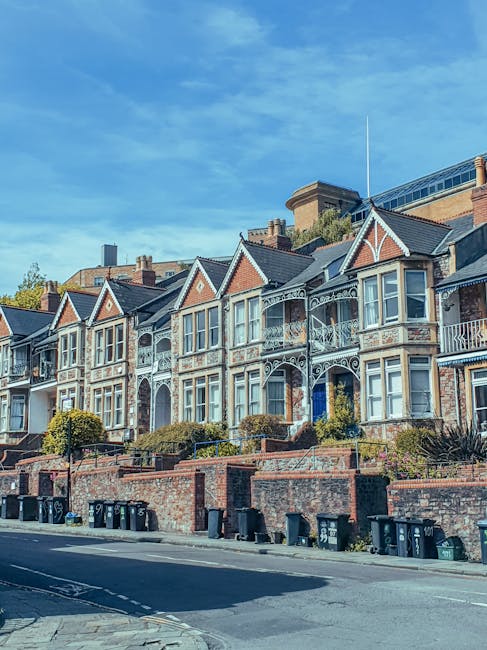 Photograph of a row of Victorian-style terraced houses in Shirley, featuring bay windows, decorative ironwork balconies, and brick facades. The houses are positioned along a street with a brick retaining wall and a sidewalk in the foreground. Multiple black wheelie bins are lined up along the curb, and the sky above is clear with a few wispy clouds. This setting illustrates typical residential architecture suitable for house removals and packing and moving services, as provided by Man with Van Shirley, supporting efficient and careful home relocation in the area.