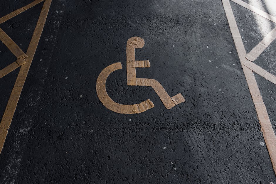 A close-up view of a designated accessible parking space on a black asphalt surface, featuring a yellow-painted wheelchair symbol with detailed lines and curves. The space is located outside a propert