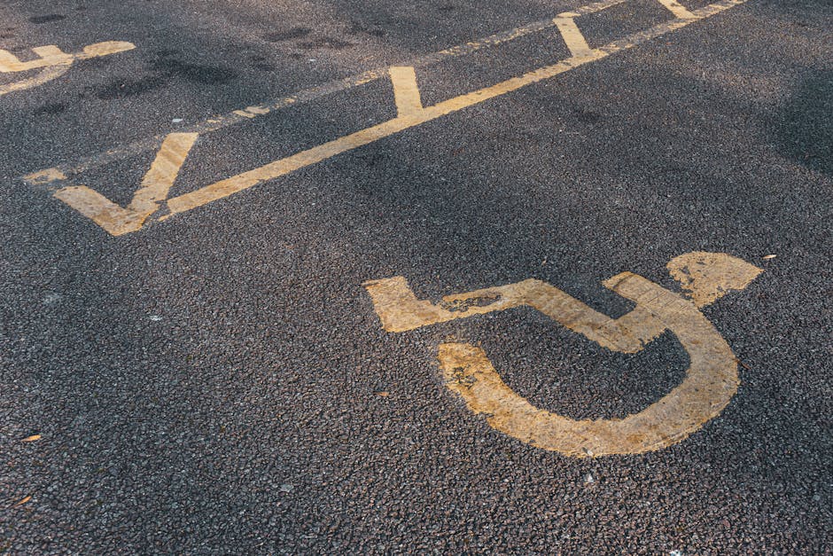 A close-up view of a designated accessible parking space on a black asphalt surface, featuring a yellow-painted wheelchair symbol with detailed lines and curves. The space is located outside a propert