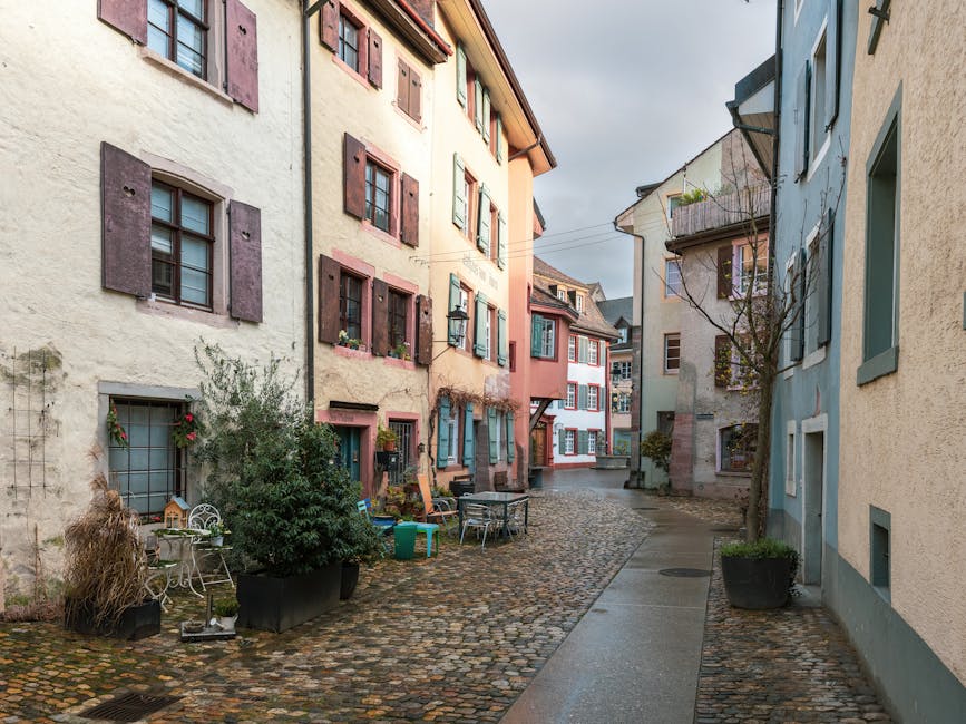 A narrow residential street with cobblestone paving, flanked by multi-story houses painted in pastel shades with contrasting window shutters. In the foreground, there are outdoor furniture and potted plants placed on the pavement, indicating a lived-in appearance. The street appears to be part of a home relocation or moving process, with no visible vehicles or moving equipment present on this portion. The surrounding environment is illuminated by soft natural light, and the environment looks calm and quiet. This setting reflects the typical urban layout where house removals services, such as those provided by Man with Van Shirley, might encounter narrow streets when transporting furniture and household items during packing and moving activities. The image emphasizes the spatial constraints and the need for careful planning when moving bulky furniture along Shirley's narrow streets.