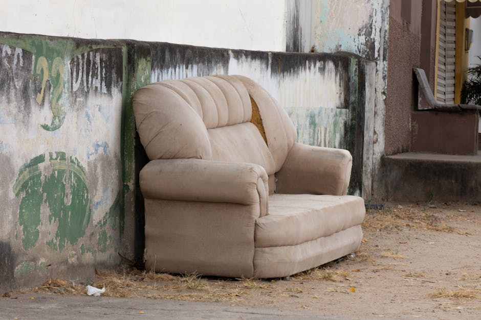 A beige upholstered armchair with padded armrests and a tufted backrest, showing visible wear and minor damage, is placed on a dirt and gravel surface outdoors, next to a weathered concrete wall with fading paint and graffiti. The wall has patches of peeling paint and discoloration, indicating exposure to the elements. Behind the armchair, there is a staircase leading to a doorway with a partially open wooden shutter. Nearby, a small piece of white paper or cloth rests on the ground. This setting suggests the piece of furniture may be in the process of being prepared for removal or relocation as part of a home moving or furniture transport service. Man with Van Shirley, a professional service specializing in removals, often manages such items during home relocation, including handling furniture lifting, loading into vans, and transport across streets like Shirley's narrow streets, as indicated by the webpage titled 'Can Bulky Furniture Be Moved on Shirley's Narrow Streets?'
