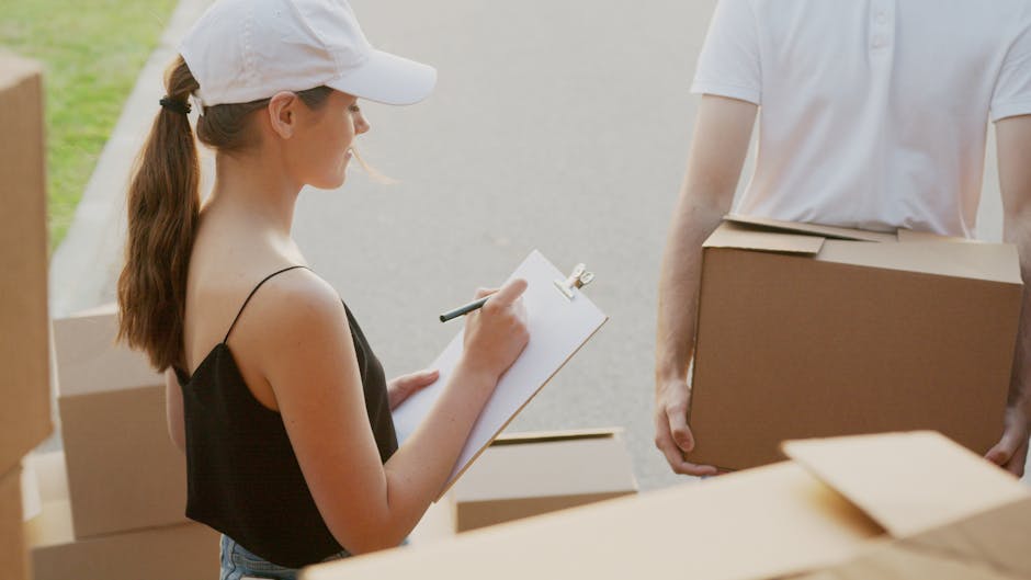 A young woman wearing a white cap and black sleeveless top is outdoors, holding a clipboard and a pen, taking notes during a home relocation; in the background, an individual in a white T-shirt is partially visible, carrying a cardboard box, with several other boxes nearby. The scene is set on a paved area adjacent to a residential property, with natural daylight illuminating the scene. The boxes are made of cardboard, some wrapped with plastic or fabric padding, indicative of furniture packing and moving preparations, consistent with house removals and furniture transport activities. This image, associated with Man with Van Shirley, illustrates the loading process typical of professional removals services, reflecting ongoing packing, planning, and logistical coordination for house moves, aligning with content about local permit requirements for Shirley removals.