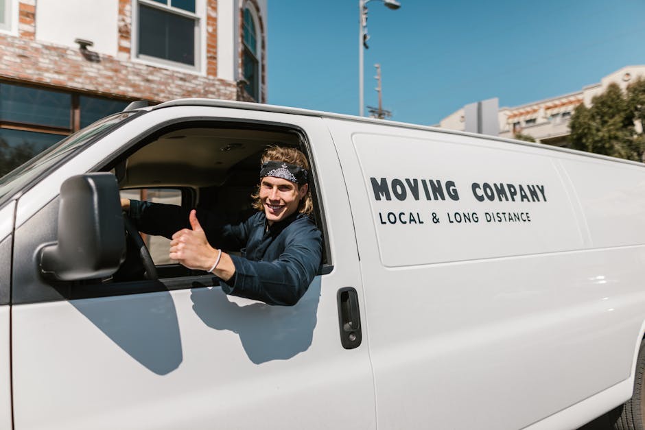 A young man with shoulder-length hair, wearing a patterned headband and a dark long-sleeved shirt, is sitting in the driver’s seat of a white moving van with 'MOVING COMPANY LOCAL & LONG DISTANCE' printed on the side. He is smiling and giving a thumbs-up gesture through the open window, indicating a positive attitude towards the home relocation or furniture transport process. The van is parked outdoors in a suburban area near Shirley Station, with buildings, signage, a utility pole, and clear blue sky visible in the background. The interior of the van shows some packing materials, including cardboard boxes and protective blankets, suggesting an ongoing loading or packing phase for a professional removal service, as provided by Man with Van Shirley. The scene captures the practical aspect of packing and loading items in preparation for a house move, emphasizing the company's local and long-distance moving services.