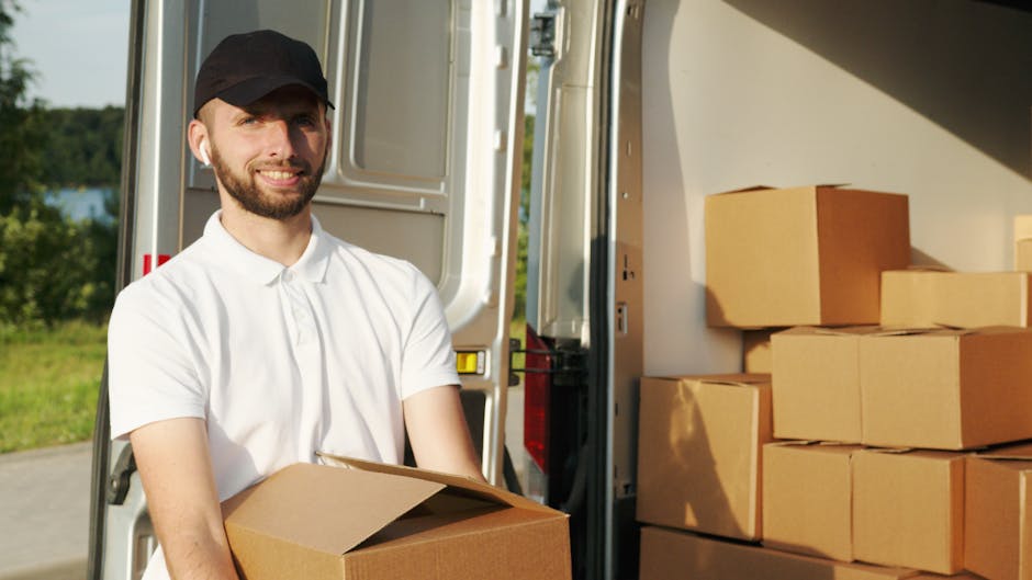 A man with a beard and wearing a black cap and white polo shirt is standing outside a moving van, holding a cardboard box in his hands. The back doors of the van are open, revealing several neatly stacked cardboard boxes inside, some wrapped in plastic and others placed directly on the floor. Outside, there is a grassy area with trees and a glimpse of water in the background, indicating an outdoor loading or unloading location. The scene is illuminated by natural daylight, and the man is preparing for a home relocation process. This image relates to furniture transport and packing procedures typical of house removals services offered by Man with Van Shirley, maintaining a professional and neutral tone suited for content regarding local moving and moving logistics.