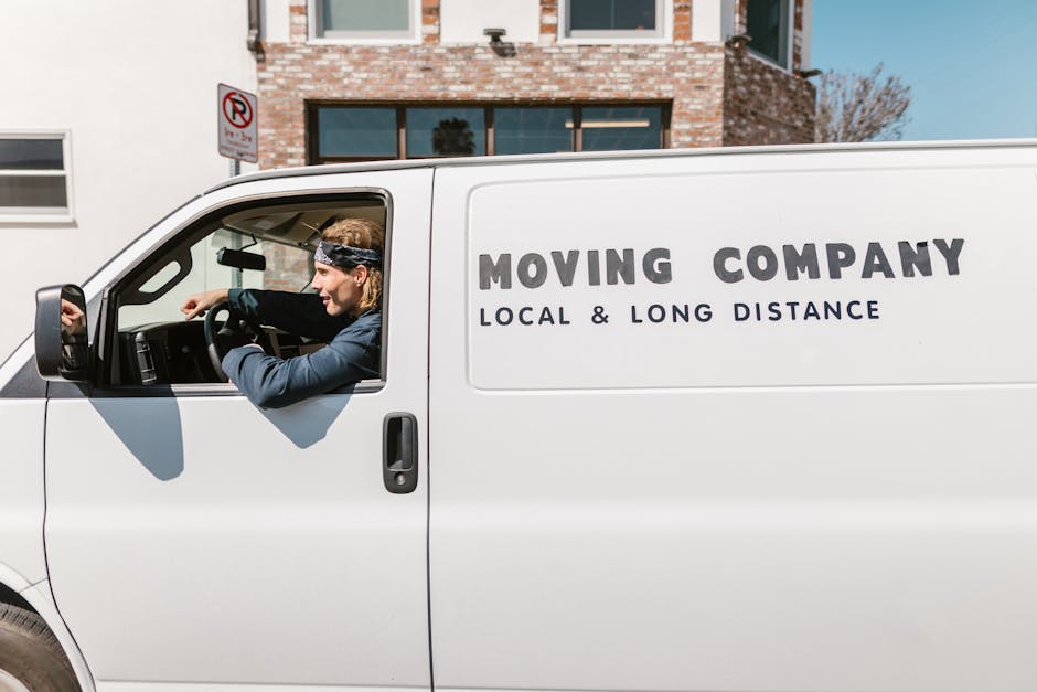 A young man with shoulder-length hair, wearing a patterned headband and a dark long-sleeved shirt, is sitting in the driver’s seat of a white moving van with 'MOVING COMPANY LOCAL & LONG DISTANCE' printed on the side. He is smiling and giving a thumbs-up gesture through the open window, indicating a positive attitude towards the home relocation or furniture transport process. The van is parked outdoors in a suburban area near Shirley Station, with buildings, signage, a utility pole, and clear blue sky visible in the background. The interior of the van shows some packing materials, including cardboard boxes and protective blankets, suggesting an ongoing loading or packing phase for a professional removal service, as provided by Man with Van Shirley. The scene captures the practical aspect of packing and loading items in preparation for a house move, emphasizing the company's local and long-distance moving services.