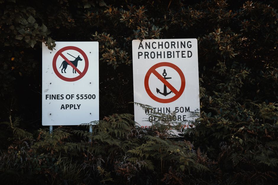 Two white outdoor signs placed among dense foliage and shrubs, with a dark background of leafy branches. The left sign displays a black silhouette of a dog crossed out with a red circle and line, alon