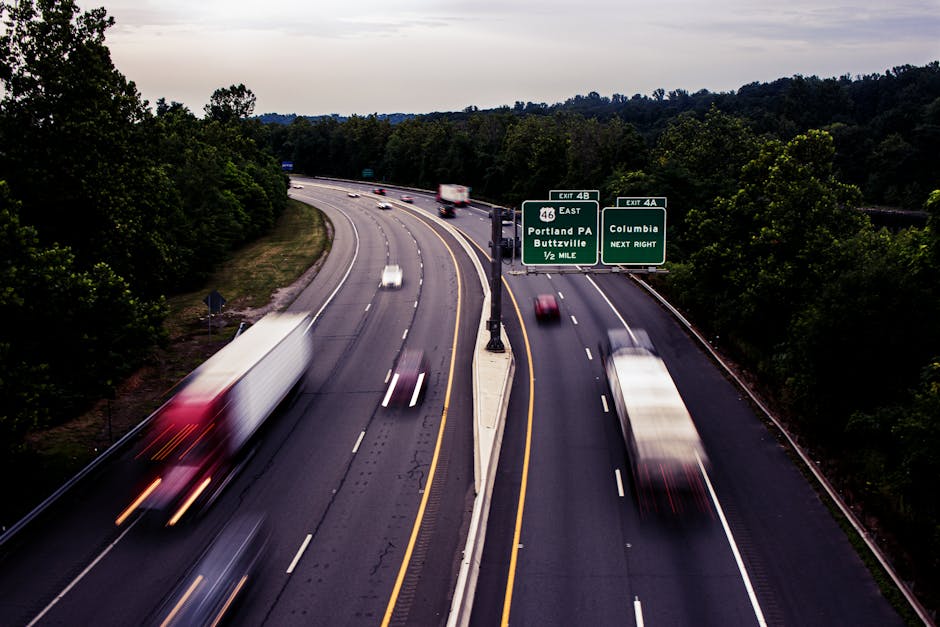 A multi-lane motorway with several vehicles, including a large white truck, a white van, and other cars, travelling in both directions. The vehicles appear blurred due to motion, indicating they are moving at high speeds. The motorway is bordered by dense green trees and bushes on either side, with overhead road signs directing towards Portland PA, Buttzville, and Columbia, as well as exit numbers for eastbound travel. The scene is captured during daylight hours, with a partly cloudy sky, and the road surface is dark asphalt with clear white and yellow lane markings. This image showcases the transportation infrastructure involved in home relocation logistics and furniture transport along busy routes, as handled by professional removals services such as Man with Van Shirley.