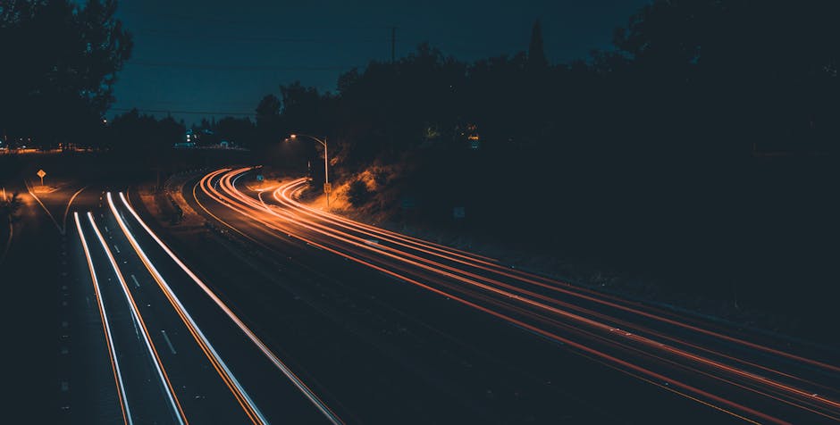 Long exposure photograph of a multi-lane road during nighttime, showing streaks of white and red light trails created by moving vehicles. The scene includes streetlights, some trees, and a dark sky in the background. The road curves gently to the left and is illuminated by the glow of nearby street lighting. This image captures the dynamic motion and steady flow of traffic, which could represent the logistical planning involved in home relocation or furniture transport services offered by Man with Van Shirley as part of their removals and moving logistics, emphasizing efficient route planning for fast moves around Shirley High Street.