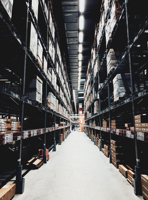 Inside a large warehouse with high metal shelving units filled with stacked cardboard boxes and packaging materials, arranged systematically along a central aisle. Bright industrial lighting illuminates the space, highlighting the neat organization of household items, appliances, and packing supplies ready for a home relocation. The concrete floor is clean and smooth, with some pallets and trolleys visible at the base of the shelves. This setting reflects the logistics involved in furniture transport and packing for local removals, supported by companies like Man with Van Shirley, who manage packing and loading processes efficiently. The scene captures the scale of storage facilities used during moves, with the perspective emphasizing the extensive inventory stored in preparation for transportation to new residences.