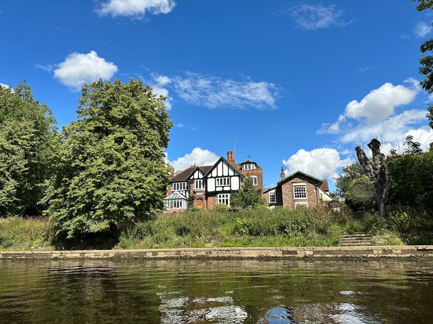 A view of a large, multi-story house with a black and white timber-framed facade and brick sections, situated on a grassy embankment next to a waterway. The house is surrounded by tall trees, including one with lush green foliage on the left and a smaller, sparser tree on the right, both positioned along a stone retaining wall with steps leading up to the property. The sky is bright blue with scattered white clouds, and the scene is illuminated by natural sunlight, highlighting the details of the house and the greenery. This setting reflects a suburban or estate area where home relocation or furniture transport might take place, with the house accessible from the water's edge, relevant to moving services provided by Man with Van Shirley.