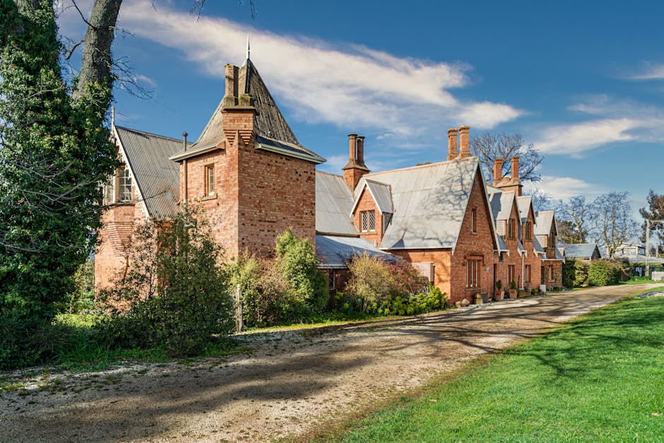 Photograph of a historic red brick house with multiple gabled roofs and tall chimneys, situated along a gravel driveway under a bright blue sky with scattered clouds. The house features traditional Tudor-style architecture with small leaded glass windows and decorative brickwork. Surrounding the property are well-maintained gardens with shrubs and flowering plants, and mature trees casting shadows on the driveway and building. Visible in the scene is part of a lawn area with neatly trimmed grass adjacent to the driveway. The image captures the exterior of the property during daytime, highlighting the structural details and natural environment, as it pertains to home relocation or furniture transport considerations for a house removal, with Man with Van Shirley providing estate-to-estate moving services.