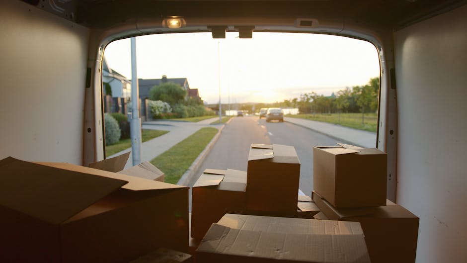The image shows the interior of a box truck used for house removals, with several cardboard boxes arranged on the floor near the open rear doors. The boxes are of various sizes, some sealed with tape and others partially open, with packing materials like paper or plastic wrap visible on some. Outside the open doors, a residential street with a sidewalk, green lawns, and houses is visible, illuminated by natural light suggests early morning or late afternoon. A few parked and moving cars can be seen on the road, indicating daytime activity. Inside the vehicle, the walls are plain and light-colored, and a loading area with no other objects or equipment present can be seen. The scene captures a moment during the packing or loading process of a home relocation, with [COMPANY_NAME] managing furniture transport and packing logistics as part of their removals services in Shirley, aligning with the webpage about urgent same-day removals.
