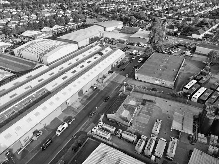 An aerial black-and-white photograph showing a commercial area with multiple industrial and warehouse buildings, some with flat roofs featuring skylights. Several parked vehicles, including vans, trucks, and cars, are visible around the premises, with a few actively being used for loading or unloading, as evidenced by the presence of open doors and materials around them. A narrow road runs through the scene with a few moving vehicles. The surroundings include residential houses, trees, and a network of streets, indicating an urban environment. During a home relocation or furniture transport process, a professional removals team such as Man with Van Shirley might carefully load packed boxes, furniture wrapped in protective materials like plastic or fabric, and appliances onto vans, coordinating the loading process within this type of industrial or warehouse setting for efficient transport to a new property.